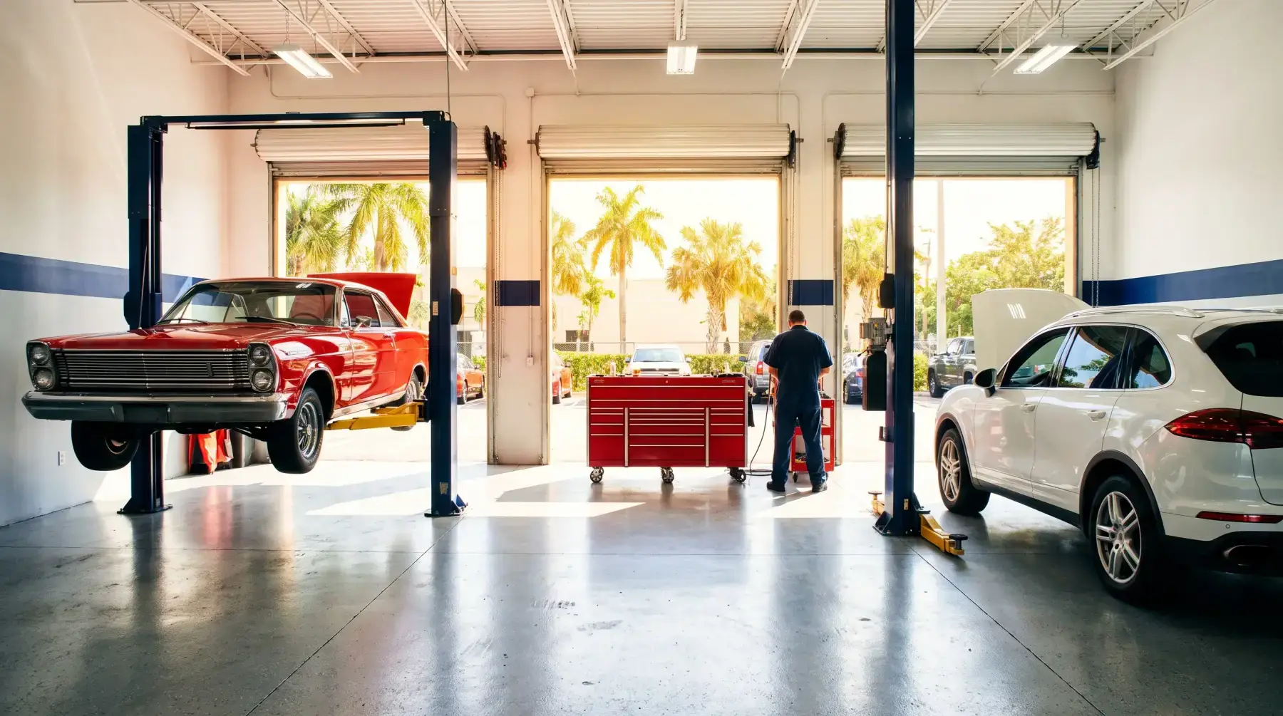 Inside the three-bay shop: a red 1960s coupe on a lift, a white SUV with its hood up, sun and palm trees through the open doors