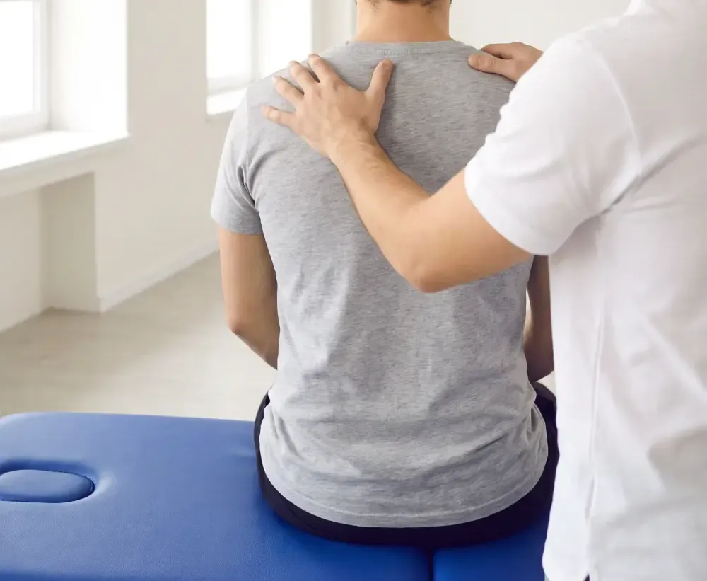 A clinician's hands on a seated patient's shoulders, seen from behind
