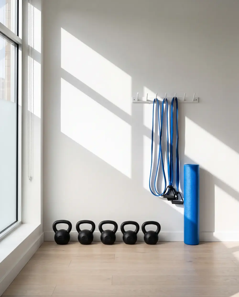 Kettlebells, blue resistance bands and a foam roller against a white wall in hard window light