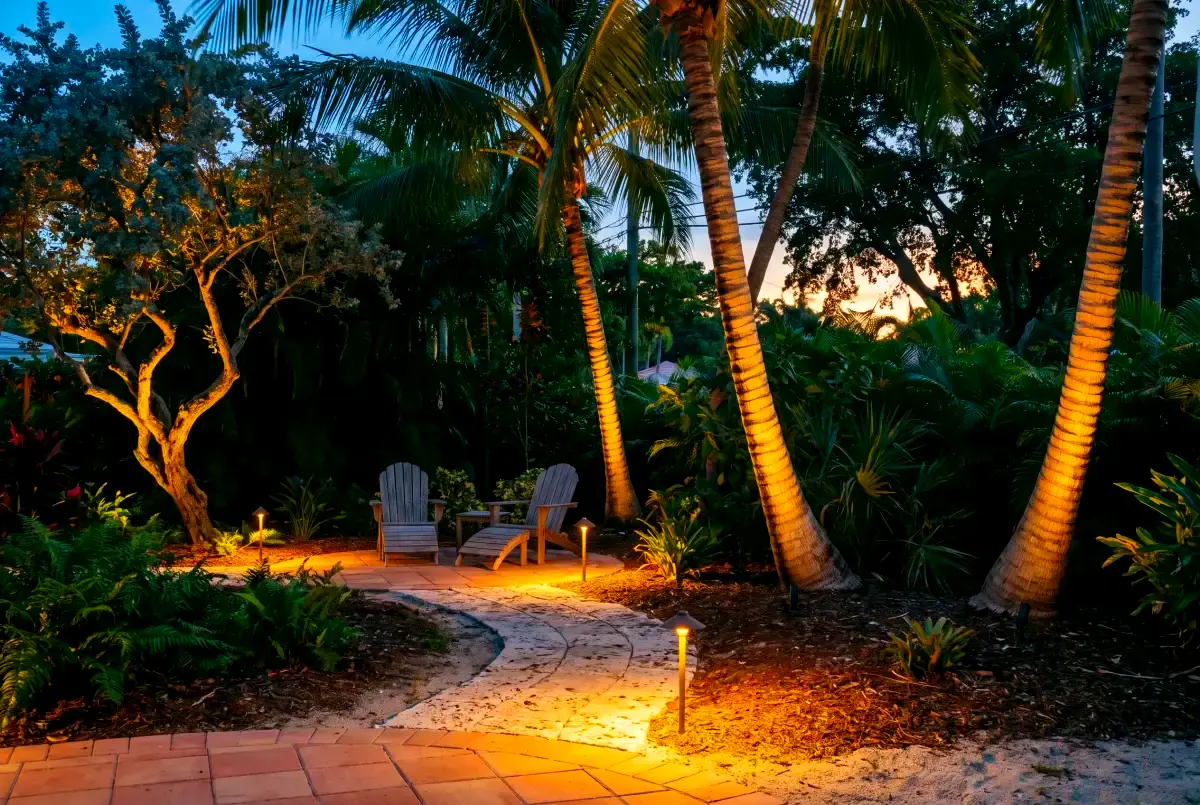 A back garden at dusk with warm path lights and uplit palm trunks