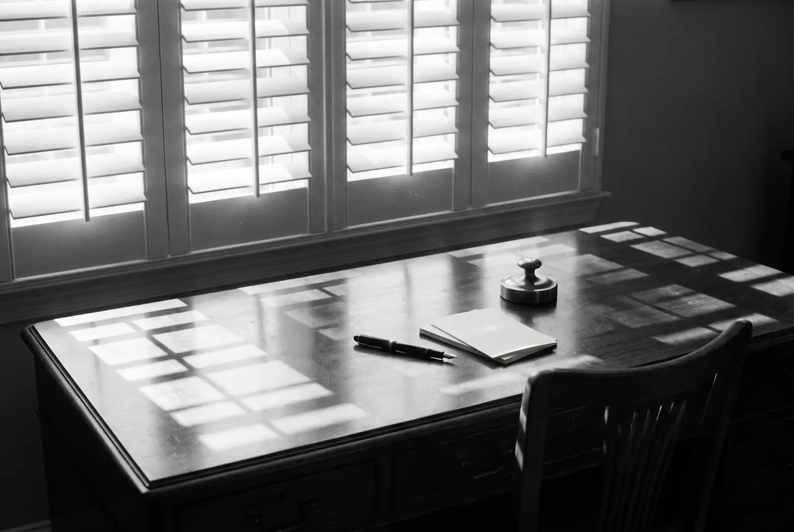 Afternoon light through plantation shutters across a bare wooden desk with a pen and paper