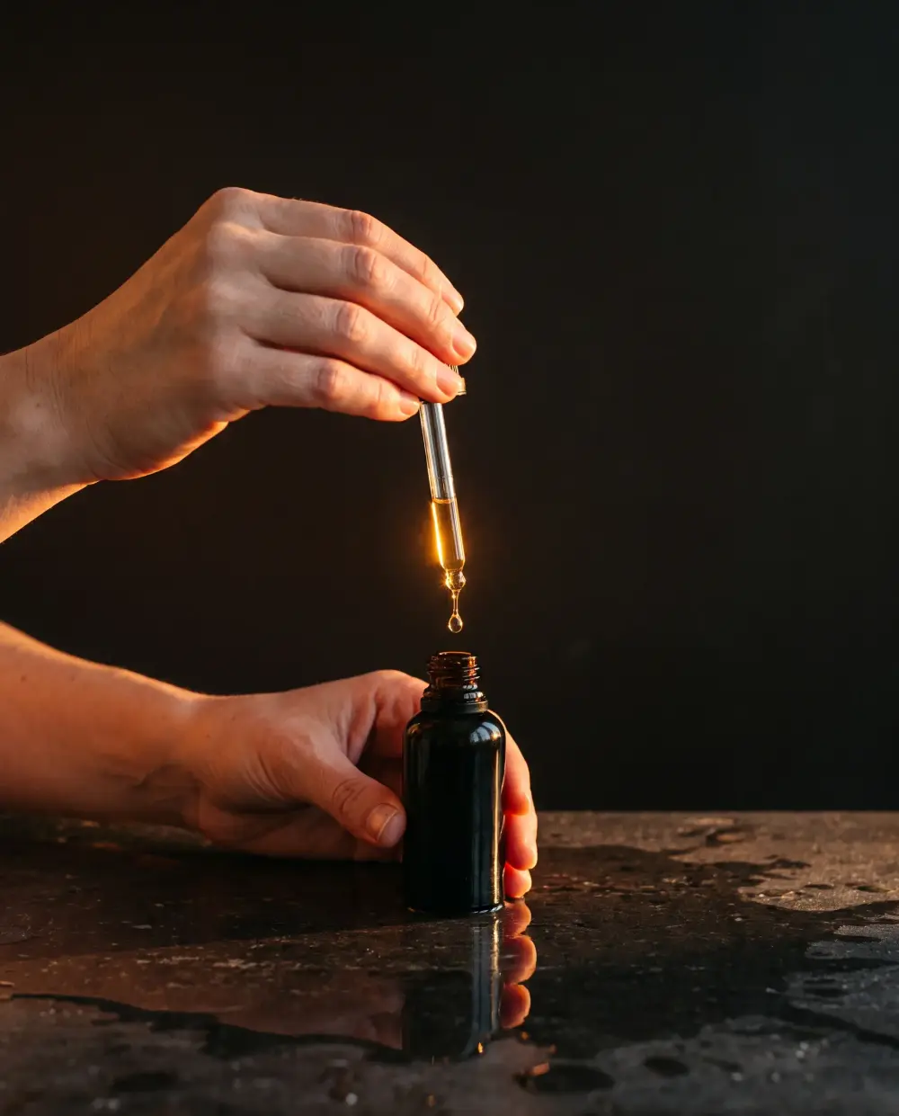 Hands drawing a glowing amber serum from a black bottle with a glass dropper, over wet black stone