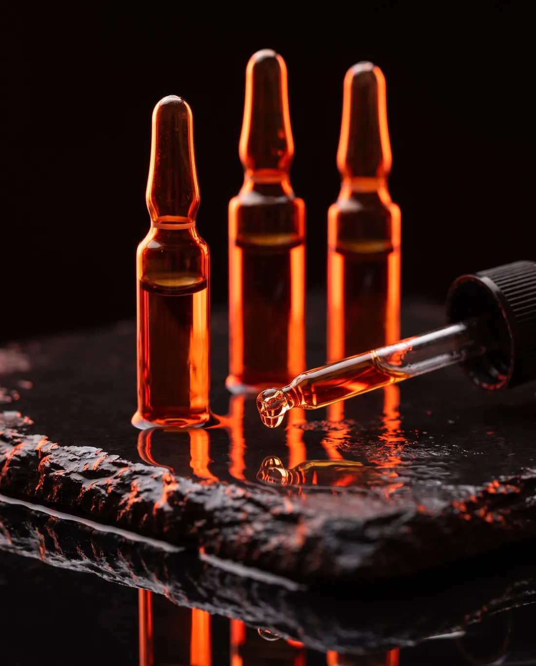 Three amber glass ampoules and a dropper on wet black stone, glowing in coral backlight