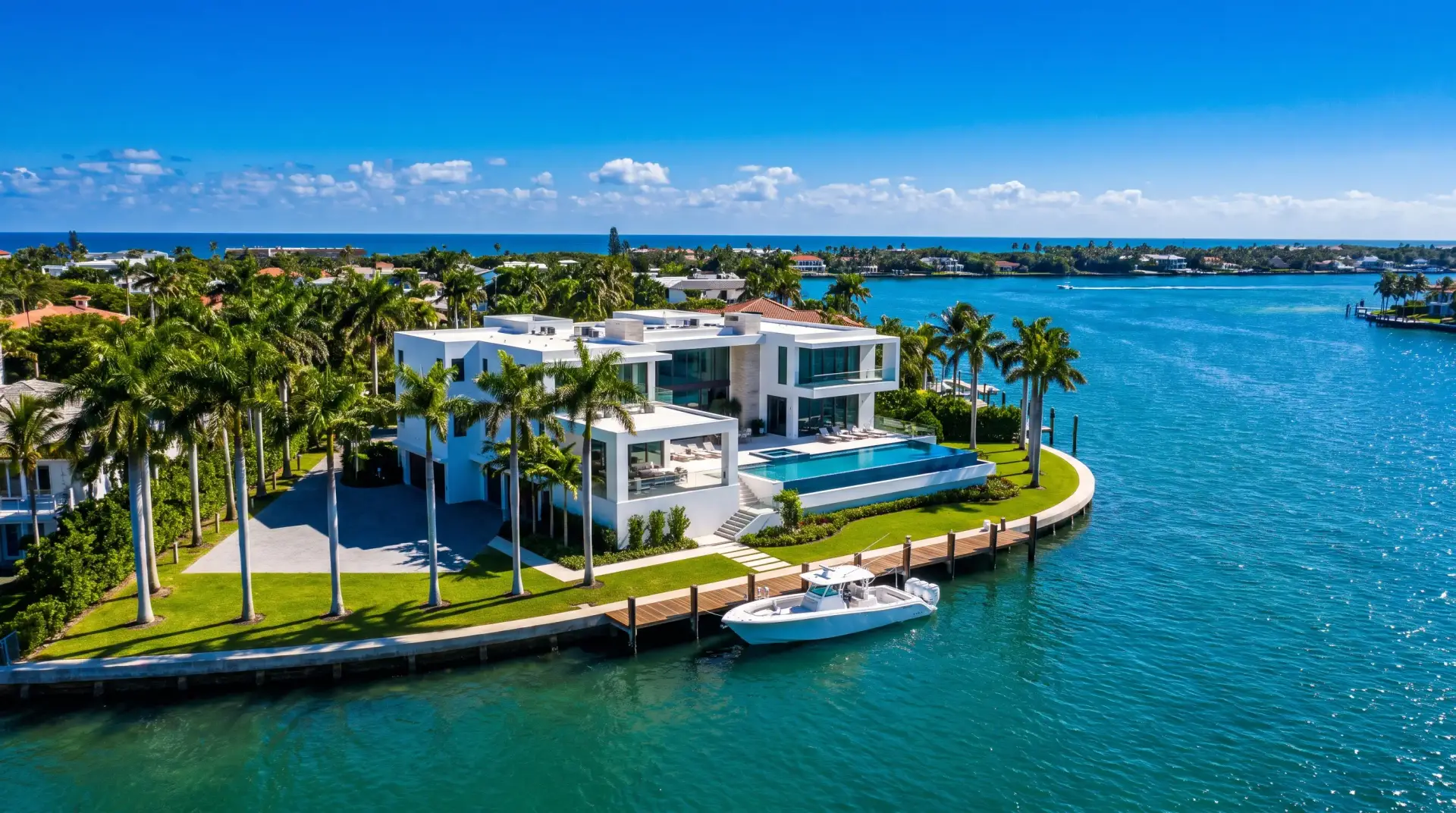 A white modern estate on a point lot, ringed by royal palms, with a boat at its dock on bright turquoise water