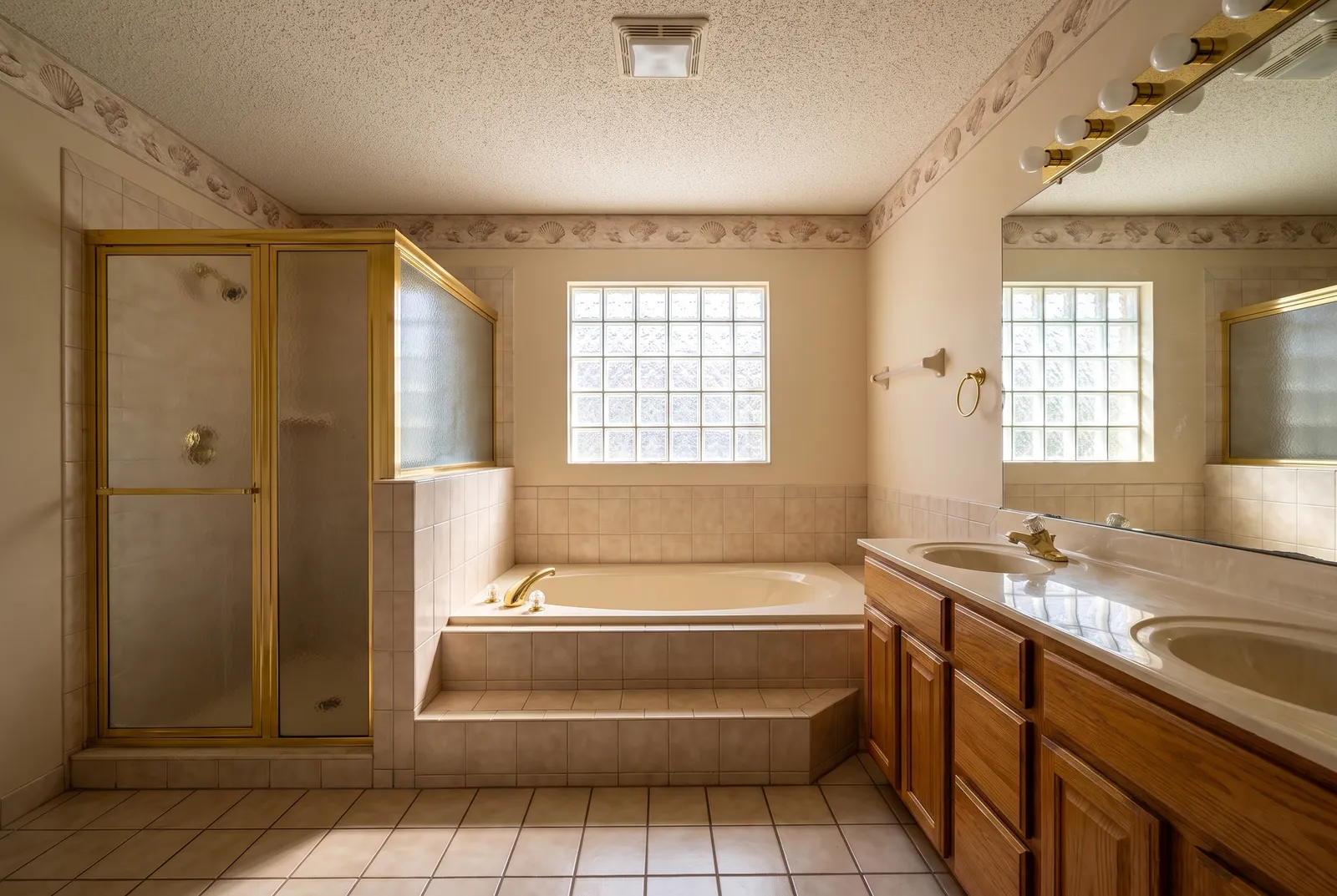 Before: the same bathroom in 1996, with a beige tiled garden tub, a gold-framed shower stall, a glass-block window and an oak vanity.