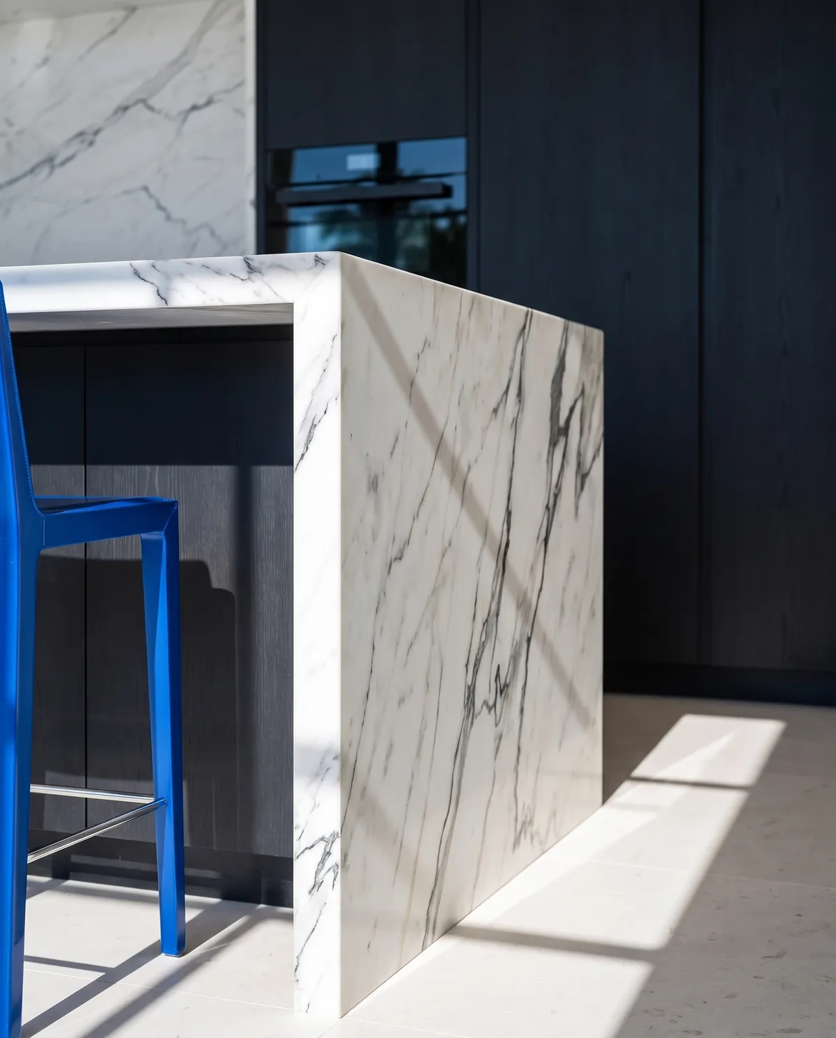 The mitred corner of a marble waterfall island where the veining runs over the edge, beside a cobalt stool.