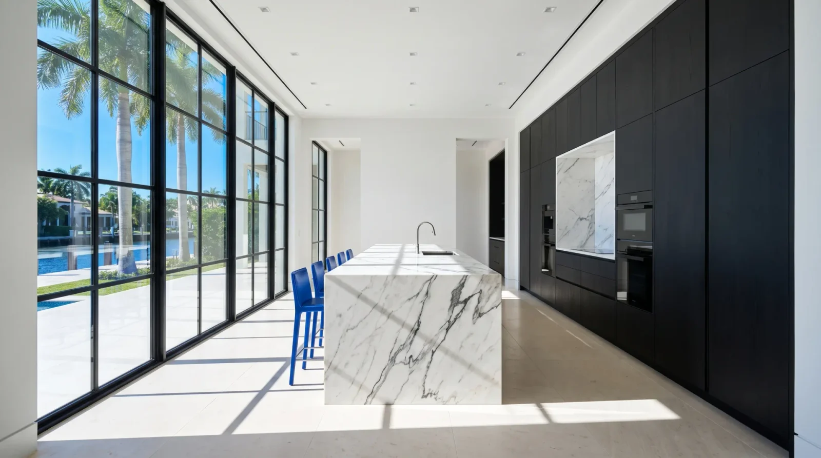 A white and charcoal kitchen with a long veined marble island, cobalt stools and a wall of steel windows onto a canal.