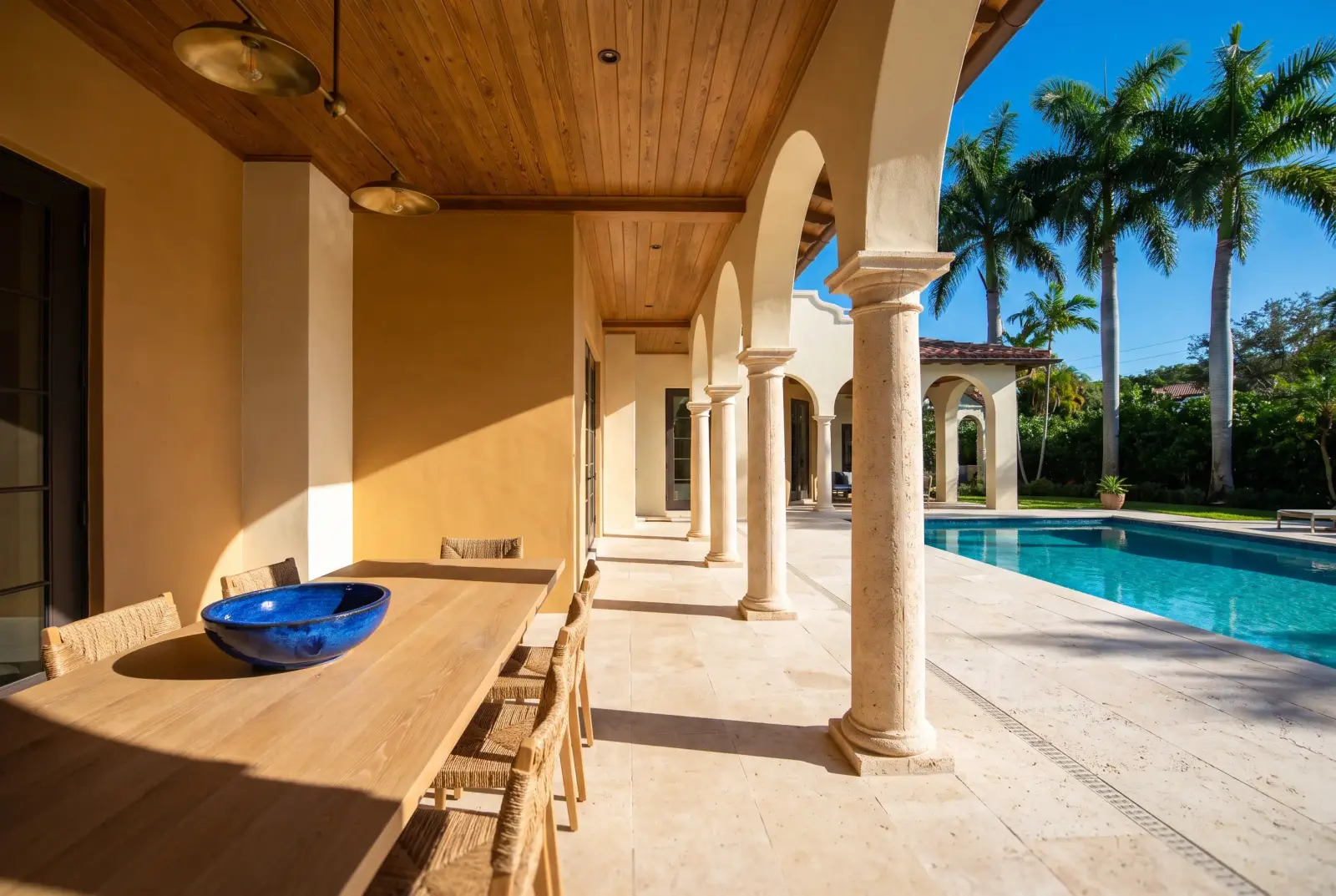 A loggia with coral stone columns and a cypress ceiling beside a long pool and royal palms under a deep blue sky.