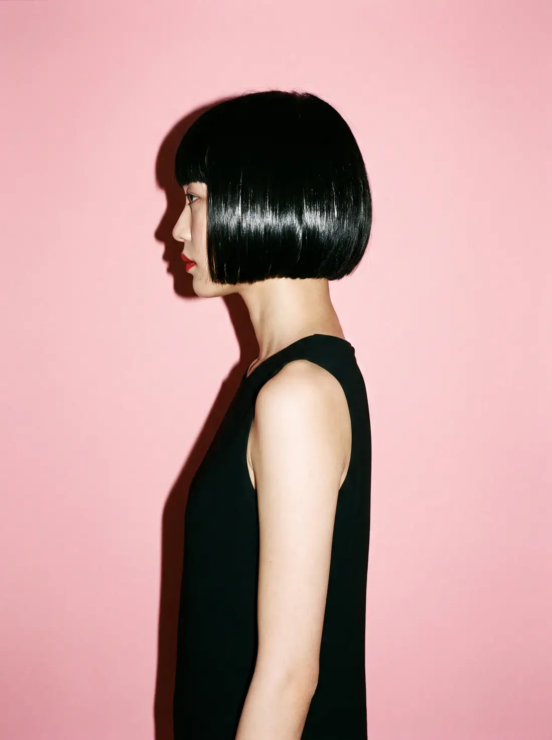 Side profile of a chin-length blunt black bob with a glassy shine against a blush backdrop.