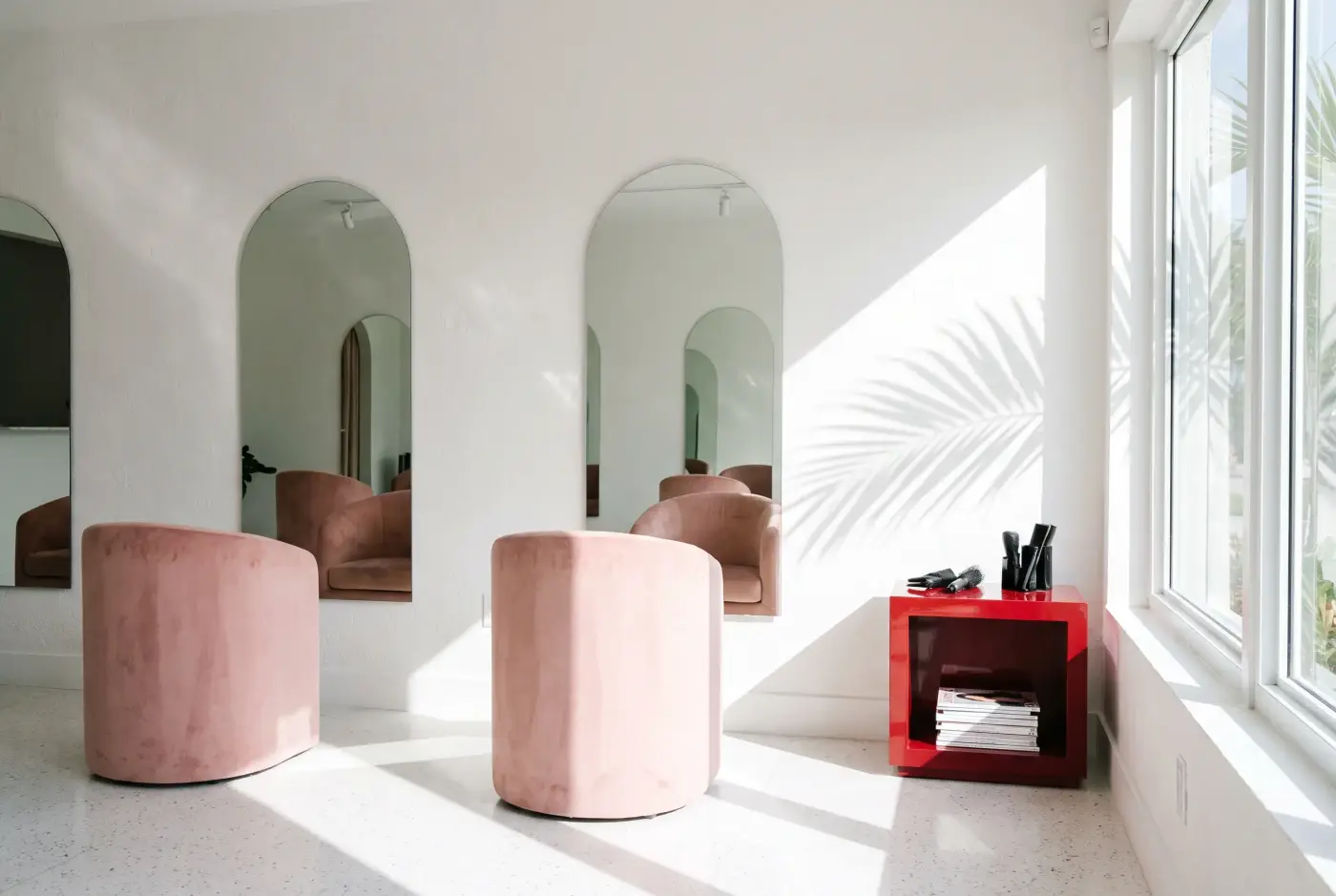 The salon interior: blush velvet chairs facing arched mirrors, white terrazzo floor and a palm shadow on the wall.