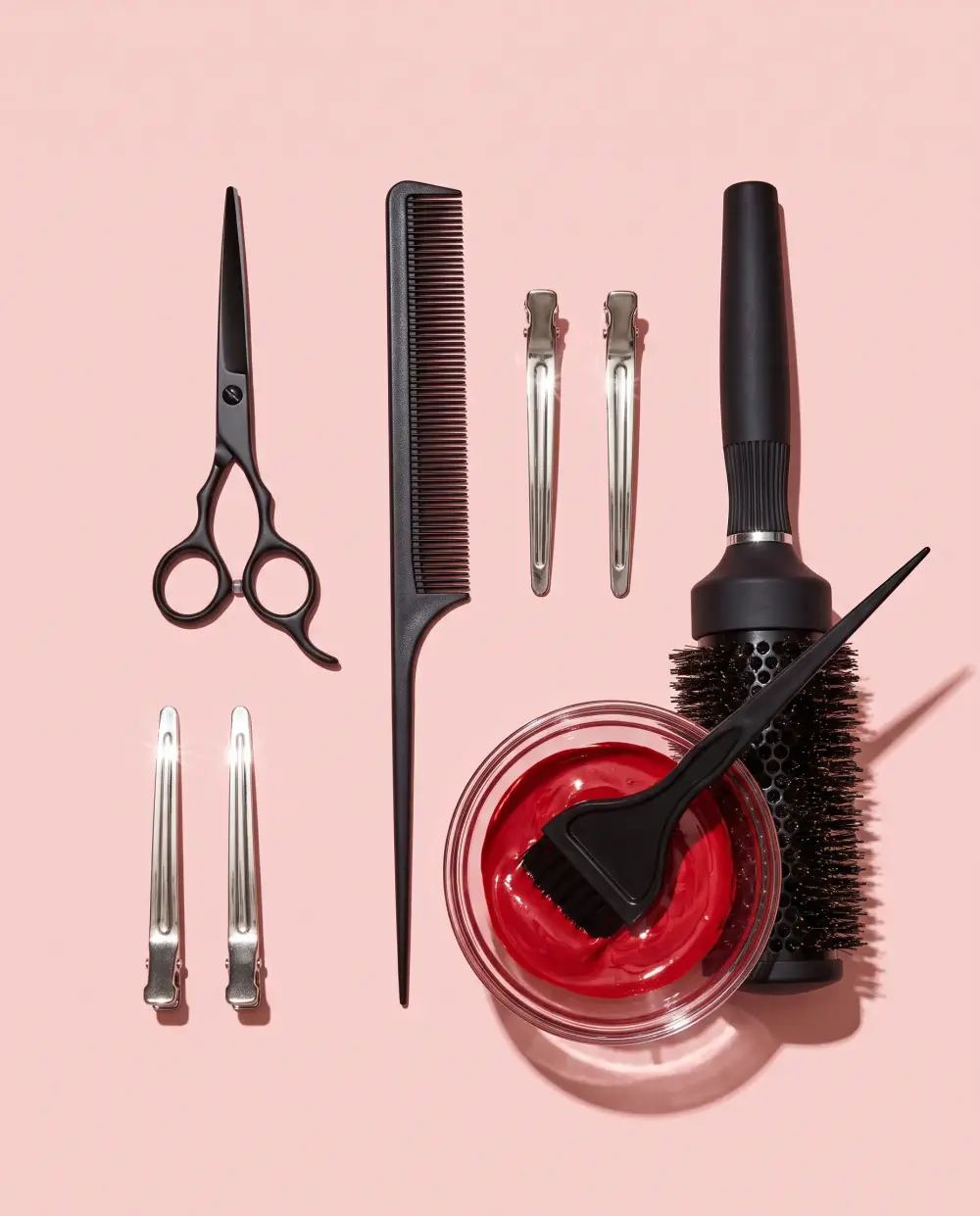 Shears, a tail comb, sectioning clips, a round brush and a bowl of red hair color laid out on blush paper.